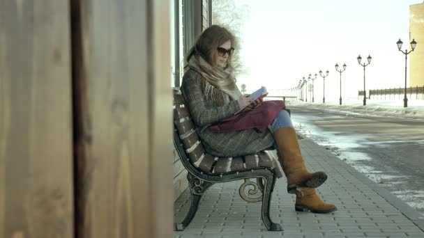 Young, beautiful, lovely woman sitting on bench in park, in hands of her notebook. She is wearing a coat. She makes notes and looks very romantic. — Stock Video