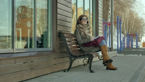 Young, beautiful, lovely woman sitting on bench in park, in hands of her notebook. She is wearing a coat. She makes notes and looks very romantic. — Stock Video