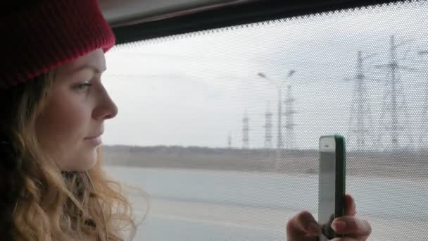 Joven mujer alegre en un sombrero rojo, viajando en autobús en un día triste. Ella toma fotos en un teléfono inteligente — Vídeo de stock