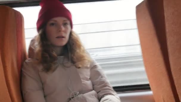 Молодая веселая женщина в красной шляпе, путешествующая на автобусе в печальный день. Она слушает музыку — стоковое видео