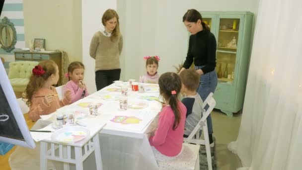 Děti chlapci a děvčata společně sedí kolem stolu v učebně a výkresu. S nimi je mladá a krásná učitelka. — Stock video