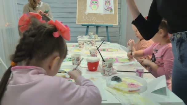 Niños niños y niñas sentados juntos alrededor de la mesa en el aula y dibujando. Con ellos está su joven y hermosa maestra . — Vídeos de Stock