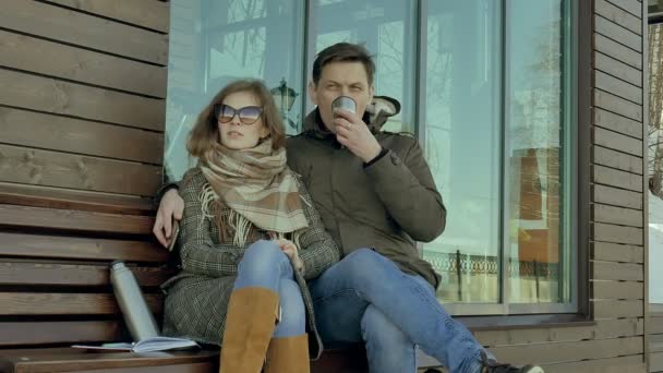 Jonge vrouw en man thee drinken uit een thermos fles in een park van de lente zittend op een bankje — Stockvideo