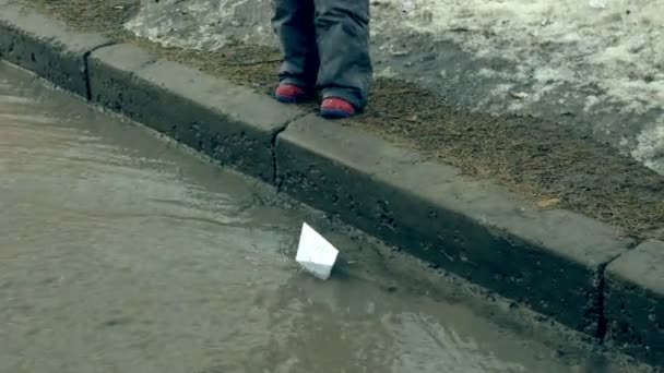 Mädchen in Gummistiefeln springt auf Pfütze nahe weißem Papierboot — Stockvideo