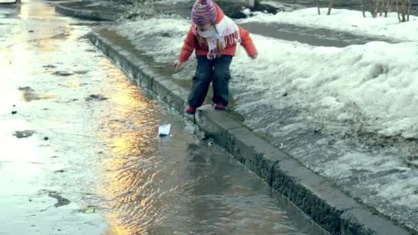 Дівчина в гумових чоботях стрибає на калюжі біля білого паперового човна — стокове відео