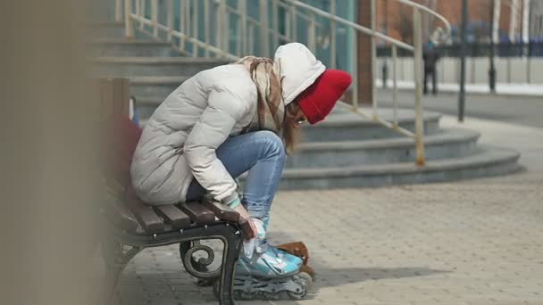 Joven mujer hermosa en ropa de abrigo deportivo y rodillos, sentado en un banco de madera y vestidos patines preparándose con patinaje — Vídeo de stock