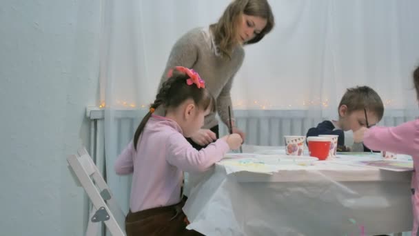 Niños niños y niñas sentados juntos alrededor de la mesa en el aula y dibujando. Con ellos está su joven y hermosa maestra . — Vídeos de Stock