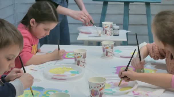 Niños niños y niñas sentados juntos alrededor de la mesa en el aula y dibujando. Con ellos está su joven y hermosa maestra . — Vídeo de stock