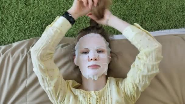 Молода жінка робить маску для обличчя з маскою для очищення, клацає на дивані зі смартфоном вдома — стокове відео