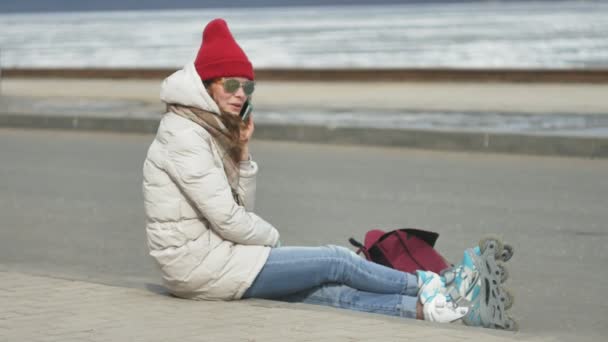 Joven hermosa mujer en un sombrero rojo con ropa de abrigo deportivo y rodillos, sentado en el camino de asfalto y hablando por teléfono — Vídeos de Stock