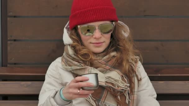 Young beautiful woman in a red hat wearing sporty warm clothes and rollers, sitting on a wooden bench drinking tea from a thermos — Stock Video