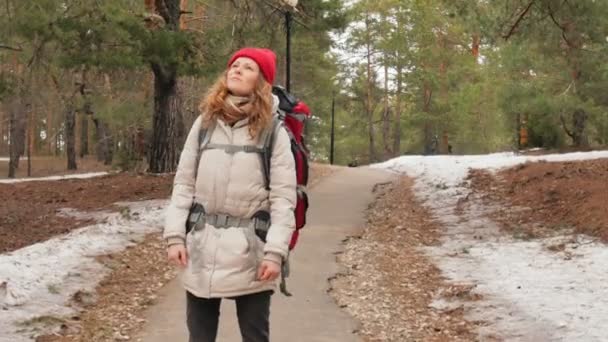 Eine attraktive junge Frau mit rotem Hut spaziert im zeitigen Frühling mit einem großen Touristenrucksack durch den Wald — Stockvideo