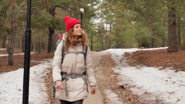 Una atractiva joven con un sombrero rojo camina por el bosque a principios de primavera con una gran mochila turística — Vídeo de stock