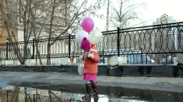 Kleines hübsches Mädchen springt nach Regen mit bunten Luftballons auf Pfützen — Stockvideo