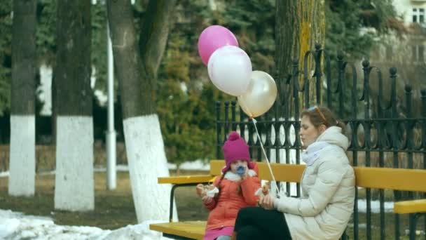 Uma menina bonita e sua mãe estão sentados em um banco de parque e comer uma deliciosa sobremesa e beber chá de uma garrafa térmica no início da primavera — Vídeo de Stock