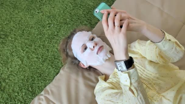 Mujer joven haciendo máscara facial con máscara de limpieza, clics en el sofá con teléfono inteligente en casa — Vídeos de Stock