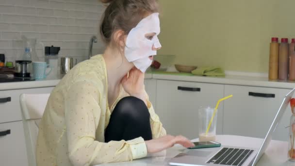 Young woman doing facial mask mask with cleansing mask, working behind laptop at home — Stock Video