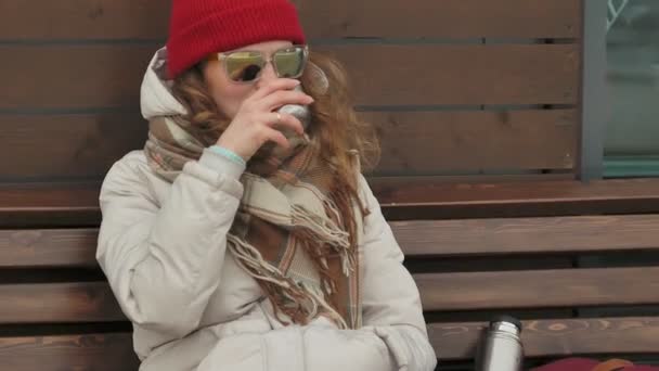 Молодая красивая женщина в красной шляпе в спортивной теплой одежде и роликах, сидящая на деревянной скамейке и пьющая чай из термоса — стоковое видео