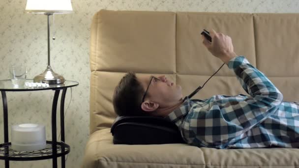 Man leży na kanapie w domu, pod głowicę do masażu, zrelaksowany stan — Wideo stockowe