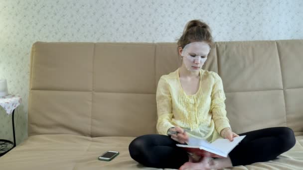 Ung kvinna gör ansiktsmask mask med rengörande mask, klickar på soffan med smartphone hemma — Stockvideo