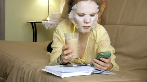 Young woman doing facial mask mask with cleansing mask, clicks on couch with smartphone at home — Stock Video