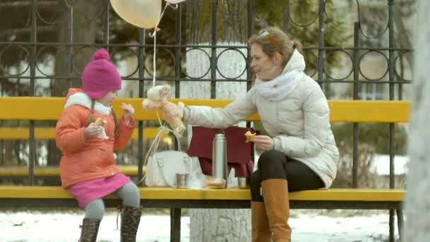 Uma menina bonita e sua mãe estão sentados em um banco de parque e comer uma deliciosa sobremesa e beber chá de uma garrafa térmica no início da primavera — Vídeo de Stock