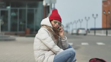 Kırmızı bir şapka giyen sportif sıcak giysiler ve Silindirler, asfalt yol üzerinde oturan ve telefonda konuşurken genç güzel kadın