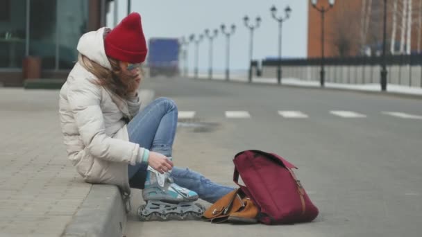 Joven hermosa mujer en un sombrero rojo con ropa de abrigo deportivo y rodillos, sentado en el camino de asfalto y hablando por teléfono — Vídeos de Stock