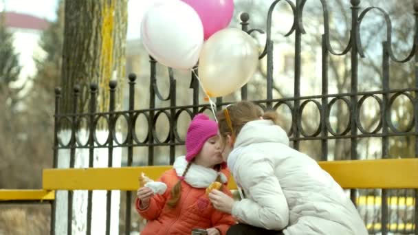 Una hermosa niña y su madre están sentadas en un banco del parque y comiendo un delicioso postre y bebiendo té de un termo a principios de primavera — Vídeos de Stock