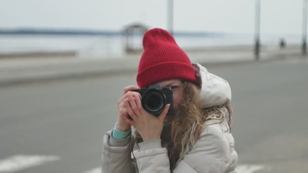 Ung vacker kvinna i röd hatt bär sportig varma kläder och rullar, sitter på asfalterad väg och tar bilder på en vintage kamera — Stockvideo