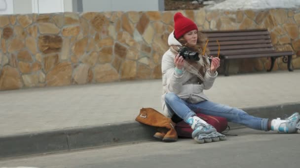 Junge schöne Frau mit rotem Hut in sportlich warmer Kleidung und Rollschuhen, die auf der Asphaltstraße sitzt und mit einer Oldtimer-Kamera Fotos macht — Stockvideo