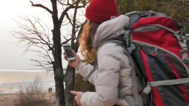 Çekici bir genç kadın kırmızı şapkalı bir büyük turistik sırt çantası ile erken baharda ormanda yürür ve telefonda fotoğraf çeker — Stok video