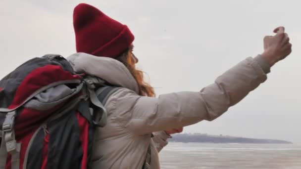 An attractive young woman in a red hat walks through the forest in early spring with a large tourist backpack and takes pictures on the phone — Stock Video