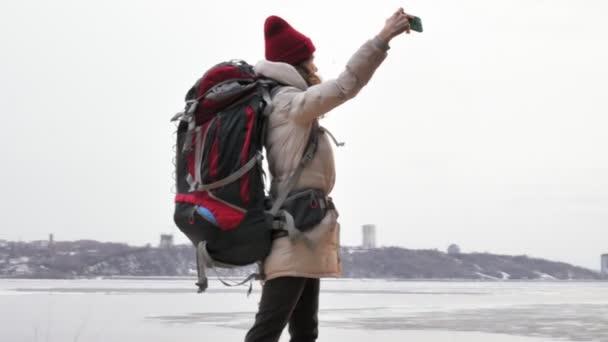 Una atractiva joven con un sombrero rojo camina por el bosque a principios de primavera con una gran mochila turística y toma fotos por teléfono — Vídeo de stock