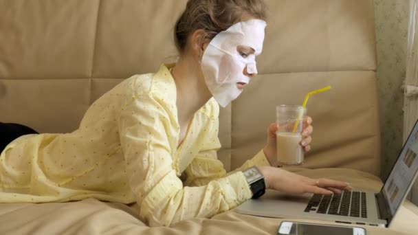 Young woman doing facial mask mask with cleansing mask, working behind laptop at home — Stock Video