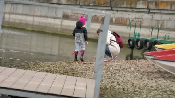 Mutter und kleine Tochter spazieren am steinigen Strand entlang, sammeln Muscheln ein und werfen Steine ins Wasser — Stockvideo