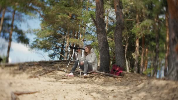 Mulher atraente olha através de um telescópio em uma floresta na margem do rio — Vídeo de Stock
