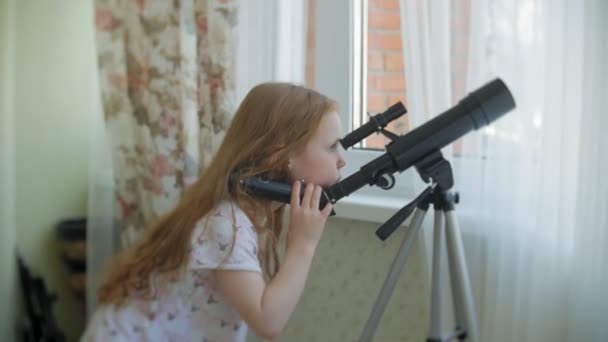 Una bambina guarda attraverso un telescopio dalla finestra della sua casa — Video Stock