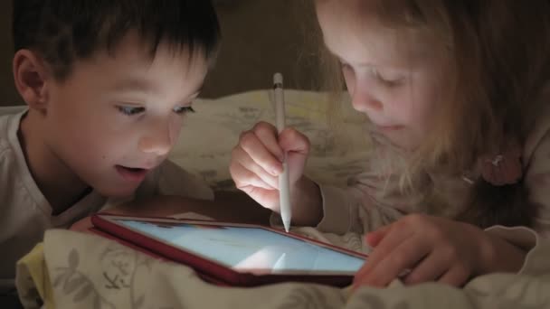 Filhinhos, irmão e irmã estão deitados na cama e brincando no tablet antes de ir para a cama . — Vídeo de Stock