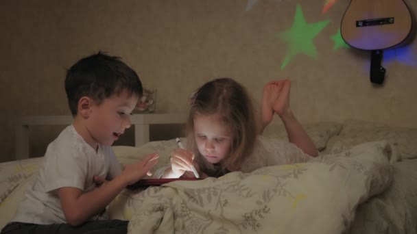 Τα μικρά παιδιά, ο αδελφός και η αδελφή είναι ξαπλωμένοι στο κρεβάτι και παίζουν στο τάμπλετ πριν πάνε για ύπνο. — Αρχείο Βίντεο