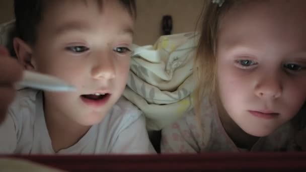 Filhinhos, irmão e irmã estão deitados na cama e brincando no tablet antes de ir para a cama . — Vídeo de Stock