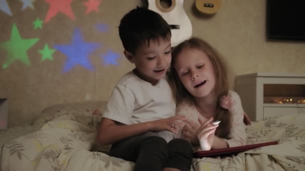 Filhinhos, irmão e irmã estão deitados na cama e brincando no tablet antes de ir para a cama . — Vídeo de Stock