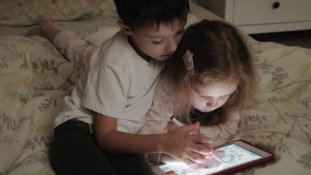 Los niños pequeños, el hermano y la hermana están acostados en la cama y jugando en la tableta antes de irse a la cama . — Vídeos de Stock