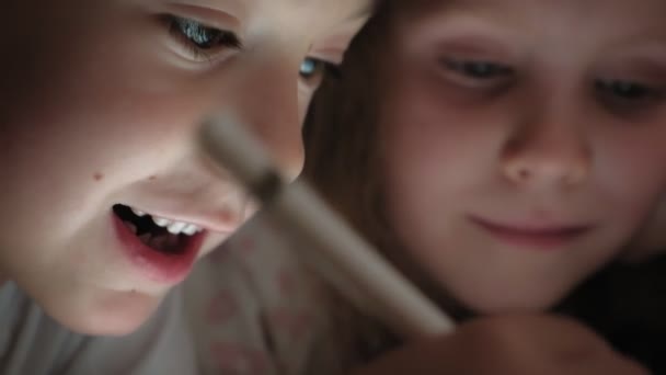 Маленькі діти, брат і сестра лежать на ліжку і грають на планшеті перед сном . — стокове відео