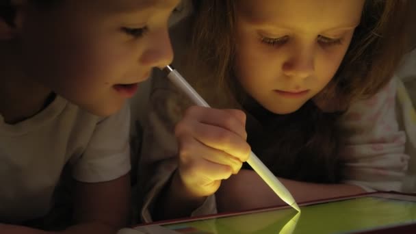 I bambini piccoli, fratello e sorella sono sdraiati sul letto e giocano sul tablet prima di andare a letto . — Video Stock