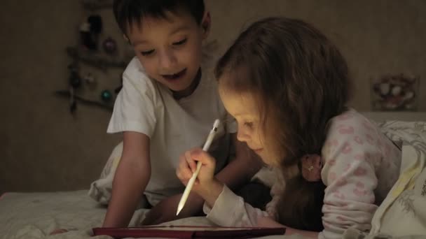 Filhinhos, irmão e irmã estão deitados na cama e brincando no tablet antes de ir para a cama . — Vídeo de Stock