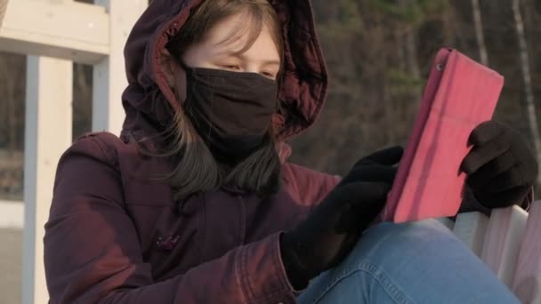 Menina em uma máscara ao ar livre usa um tablet. isolamento — Vídeo de Stock