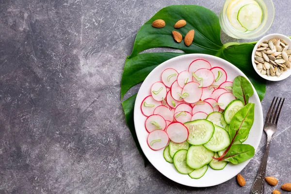 Salatalıklı hafif turp salatası, limonlu detoks içeceği, badem ve gri arka planda balkabağı çekirdeği. Sağlıklı beslenme konsepti. Üst görünüm