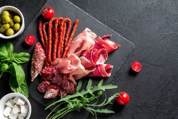 Tapas konsepti. Prosciutto, sosisli sucuk, zeytin, vişneli domates, roka, fesleğen ve peynir siyah tahtada. Antipasti konsepti. Üst görünüm.