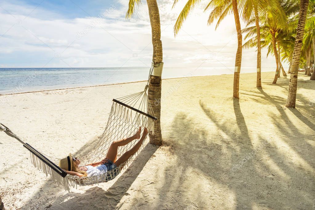Woman in hat relaxing on hammock on the beach and enjoying sunset. Travel and vacation concept.